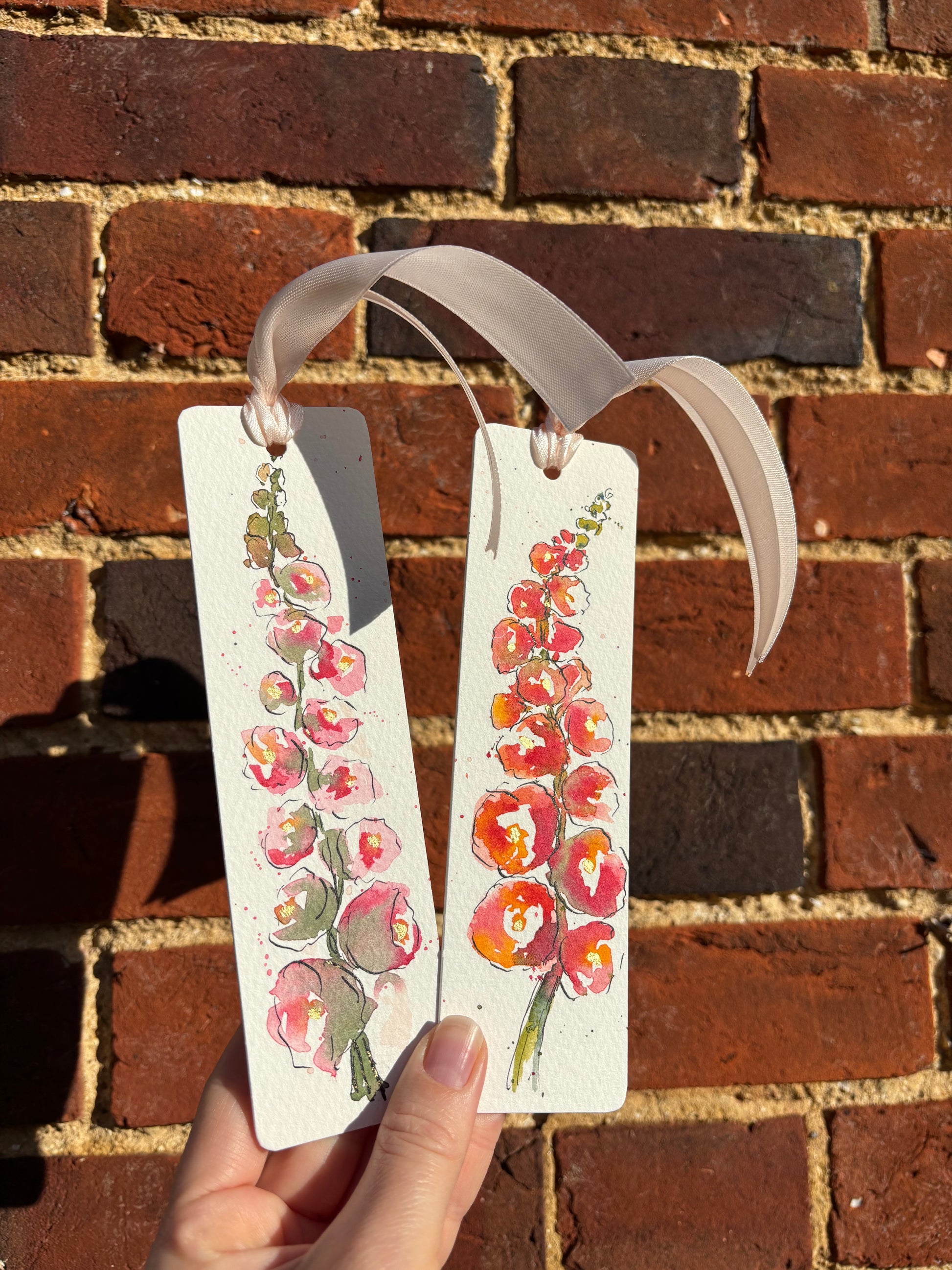 Hand holding a hollyhock and gold leaf bookmark against a brick wall background