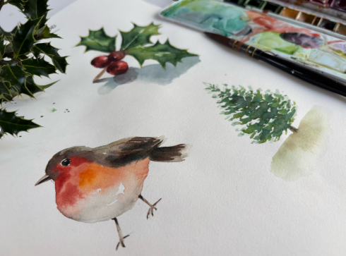 A watercolor painting of a robin bird with green holly leaves and berries, a small Christmas tree, and a pale beige shape on white paper, with a paint palette and brush partially visible in the background.