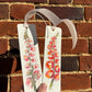 Hand holding a hollyhock and gold leaf bookmark against a brick wall background