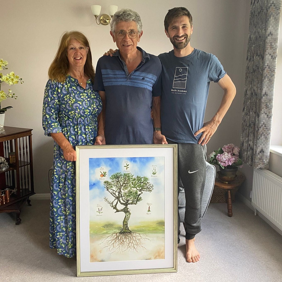 Three people standing in a living room holding a framed commissioned watercolour painting of a tree of life with botanicals,